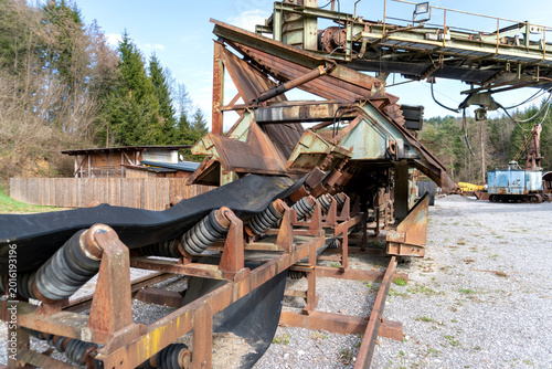 Rusted quarry conveyor belt with metal rollers, springs, rails, and heavy support structures in an outdoor industrial site. The scene shows mining equipment, material transport, mechanical engineering