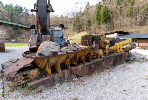 Old yellow mining conveyor loaded with rough stones and rusted chains in an outdoor quarry equipment area. The image shows raw material handling, industrial transport, heavy machinery, metal corrosion