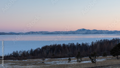 Wide winter landscape with frozen Lake Baikal in the Maloye More strait, pastel sunset sky, distant snowy mountains and forested shoreline, Siberia, Russia
