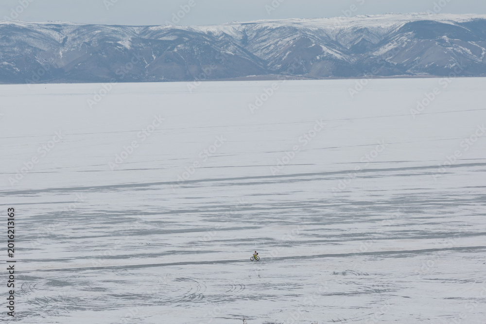 Fototapeta premium Wide winter landscape of frozen Lake Baikal with a lone cyclist riding across the ice, distant snow-covered mountains and overcast sky, Russia, Siberia