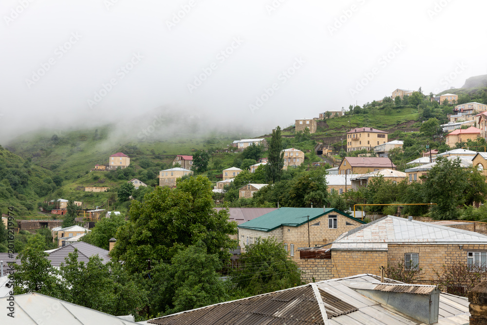 Fototapeta premium Panoramic view of Kubachi village on a green mountain slope with stone houses and metal roofs, low clouds and fog after rain, Dagestan, Russia, Caucasus