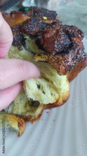 Close up view showing Caucasian hand tearing apart fluffy golden yeast dough inside traditional homemade festive bread roll covered with dark crunchy poppy seeds