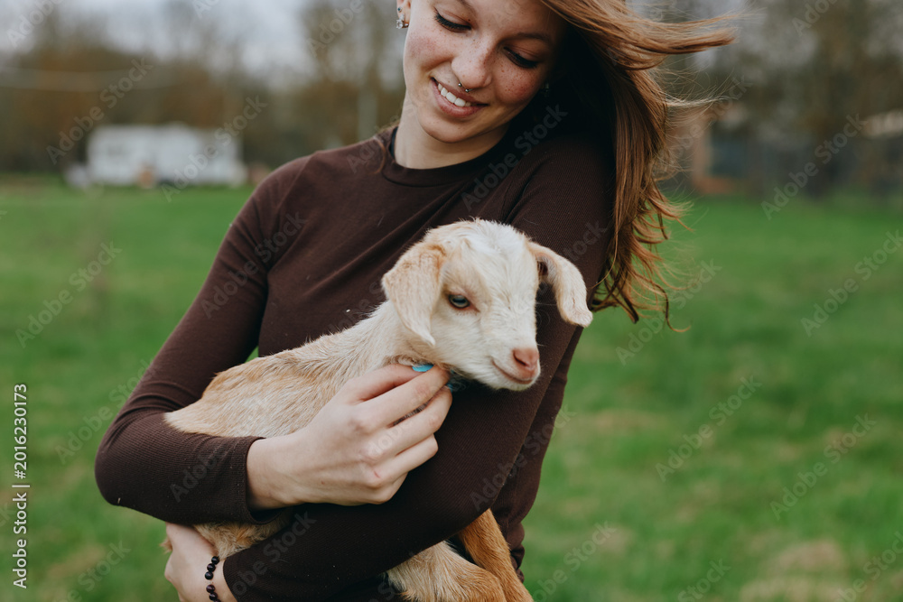 Obraz premium Smiling woman hugs a goat kid in a grassy field, highlighting a gentle countryside lifestyle, connection with animals, and pastoral calm under soft natural light, creating a warm, hopeful mood.