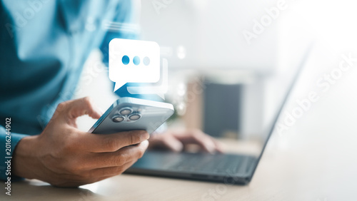 Person using smartphone beside laptop with chat icon overlay, representing online messaging, mobile communication, customer support, social media interaction, app notification, and digital connection.