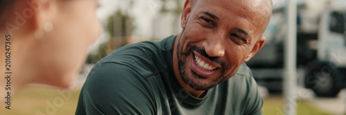 Close-up side view of people in sportswear cheerfully talking and sitting on a park bench, two multi-ethnic male and female athletes relaxing and chatting. Сouple goals and motivation. Panoramic