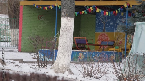 Outdoor kindergarten playground featuring decorated wooden pavilion with colorful bunting flags mural paintings miniature chairs benches basketball hoop surrounded by snow covered ground