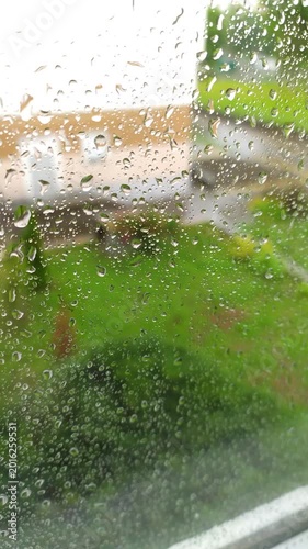 Transparent water droplets condensation sliding down window glass pane overlooking blurred green garden lawn backyard during summer rain storm.