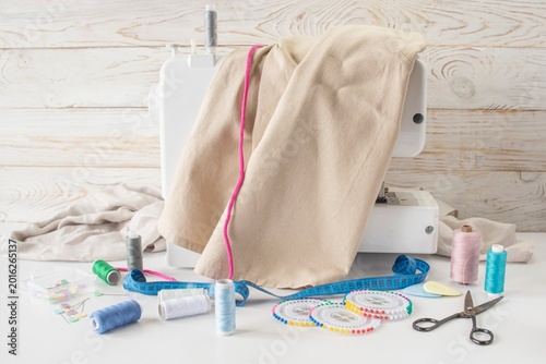 Sewing supplies on a white table with a sewing machine in the background. Sewing machine with fabric and sewing accessories on a white table.