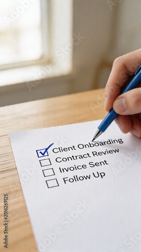 A close-up of a hand holding a blue pen checking a box on a paper checklist for client onboarding and contract review, sitting on a wooden desk near a window, conveying a sense of business management