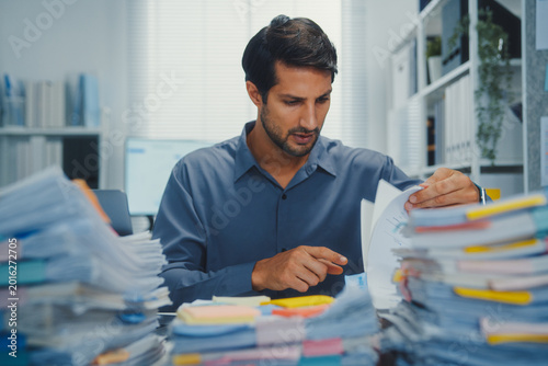Overworked young Latin businessman reading document in large stack of paperwork files at office desk. Tax audit deadline, accounting stress, corporate burnout, bookkeeping, bureaucracy concept.