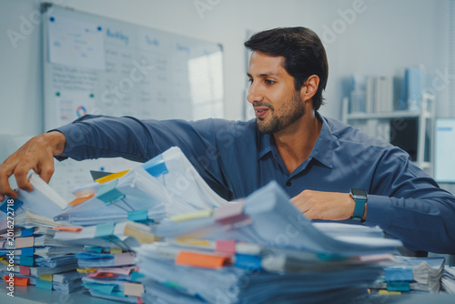 Overworked young Latin businessman reading document in large stack of paperwork files at office desk. Tax audit deadline, accounting stress, corporate burnout, bookkeeping, bureaucracy concept.