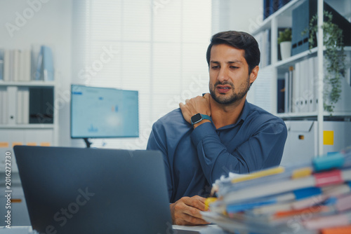 Young Latino businessman suffering shoulder pain and neck ache at office desk with computer. Office syndrome, muscle massage, overworked stress, bad posture, and health insurance injury concept.