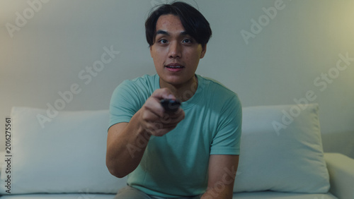 Bild auf Leinwand Young Asia male with casual cloth sit on sofa hold remote control look at television scrolling choose favorite movie chanel at living room in cozy house at night