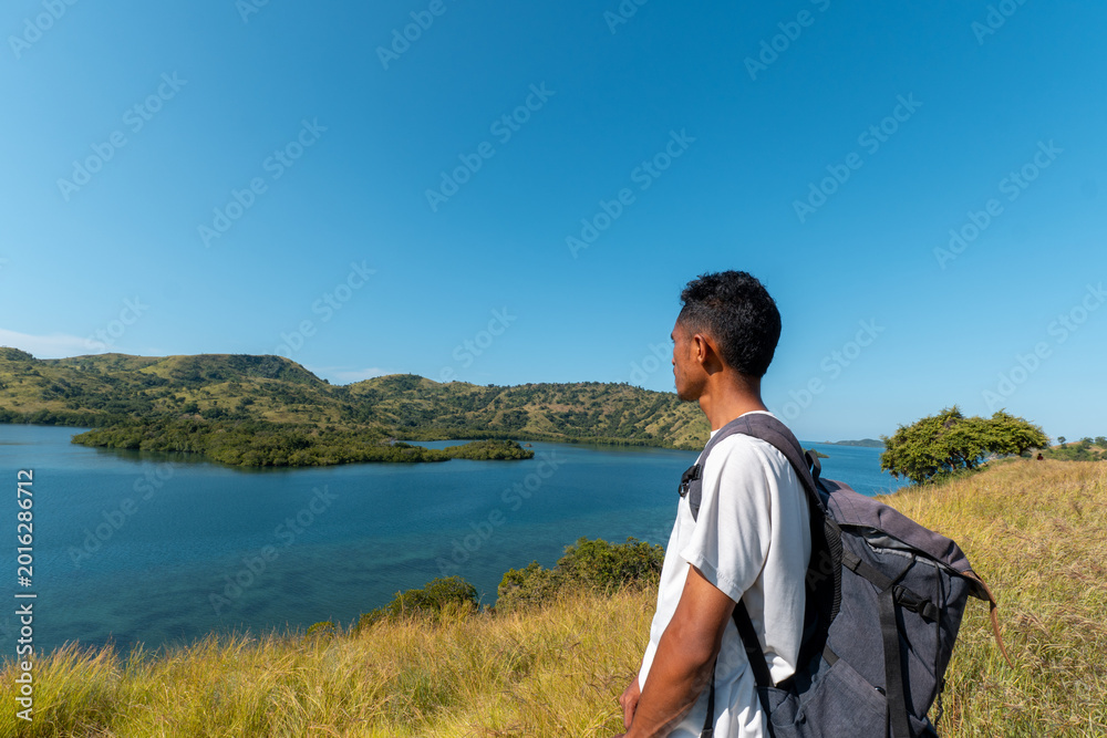 Fototapeta premium Rear view of a young Asian traveler with a backpack standing on a grassy hilltop, admiring the view of a calm blue sea bay and coastal mountains.