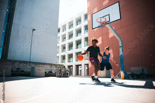 Two young women are actively playing basketball on an outdoor court. One woman dribbles the ball, moving past her opponent who is defending. They are enjoying a sunny day of sports.