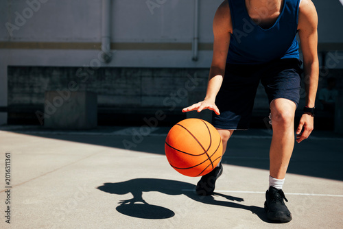 A person in athletic wear dribbles an orange basketball on a sunny outdoor court. The player's hand is above the ball, mid-dribble, showcasing active play.