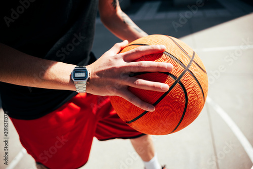 A player holds a basketball on an outdoor court, hands gripping the textured surface. A digital watch is on their wrist. This image conveys readiness and passion for the game.