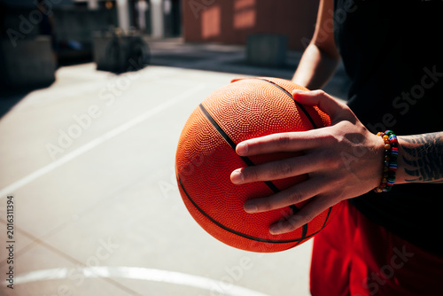 A young athlete holds a textured basketball firmly with both hands on an outdoor court. The person wears a black shirt and red shorts, showcasing a casual, active lifestyle.