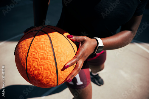 A dark-skinned woman holds an orange basketball firmly in her hands. She is dressed in athletic wear, including shorts with a number and a smartwatch, on an outdoor court.