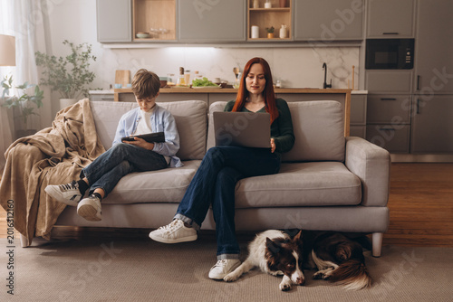 Family digital routine: woman and boy with laptop, tablet and dog in the kitchen-living room
