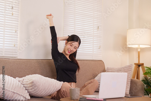 Asian woman stretching arms working from home office with laptop computer on sofa feeling happy relaxed during break time remote work freelance lifestyle