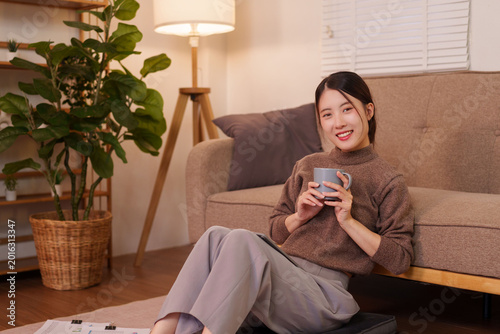 Foto Asian woman relaxing at home holding coffee mug sitting on floor by sofa in cozy