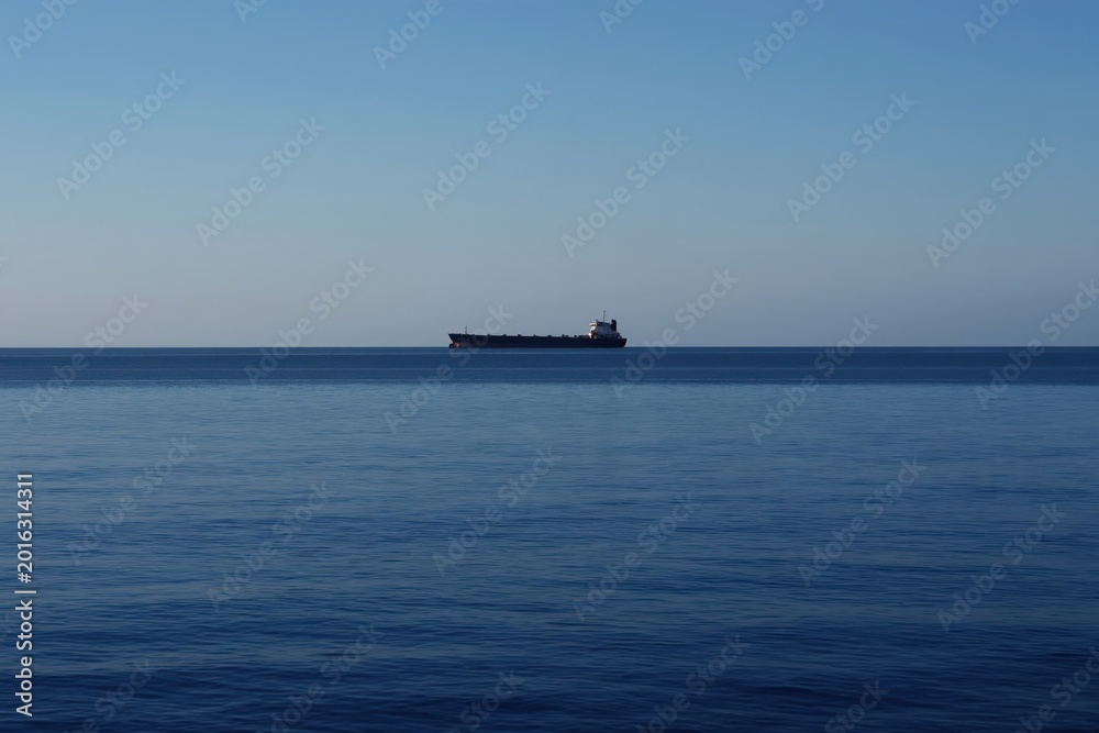 Fototapeta premium Large cargo ship sails on calm blue ocean under clear sky. The vessel travels on horizon line. Marine shipping and international trade themes.