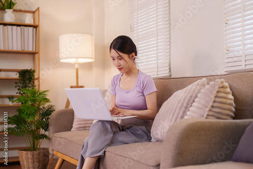 Принти на полотні young Asian woman working from home using laptop computer sitting on sofa in mod