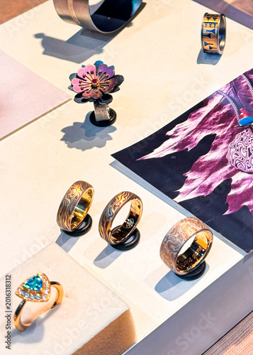 Collection of unique handcrafted rings and jewelry displayed in a Venice boutique. Includes mokume-gane bands, a flower ring and a blue gemstone piece on a neutral background.