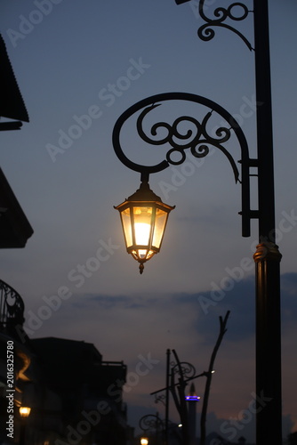 A vintage street lamp stands silhouetted against a fading blue twilight sky. Soft orange light from the lantern creates a warm focal point amidst the cooling evening atmosphere.