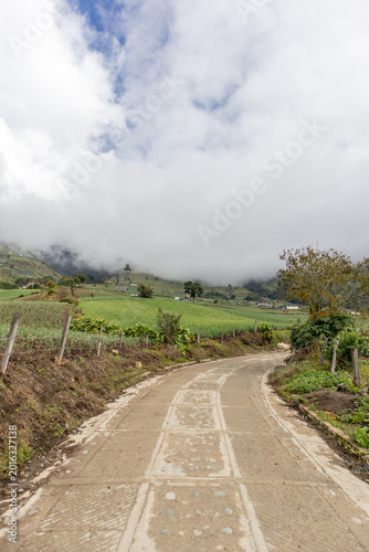 Drone aerial view of a rural farm surrounded by lush green mountains in Tenerife Valley, Colombia. Agricultural fields with neatly planted crops, countryside houses, rolling hills, and natural landsca