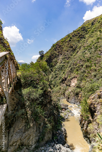 Scenic view of a narrow mountain canyon with a flowing river and an old steel bridge crossing the landscape. Rugged cliffs, native vegetation, and clear blue skies highlight the natural beauty of the 