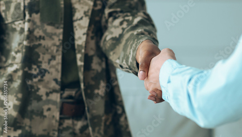 Two individuals shake hands in a friendly manner. One person wears a military uniform, while the other is in casual clothing. The setting appears to be indoors, possibly during a community event.