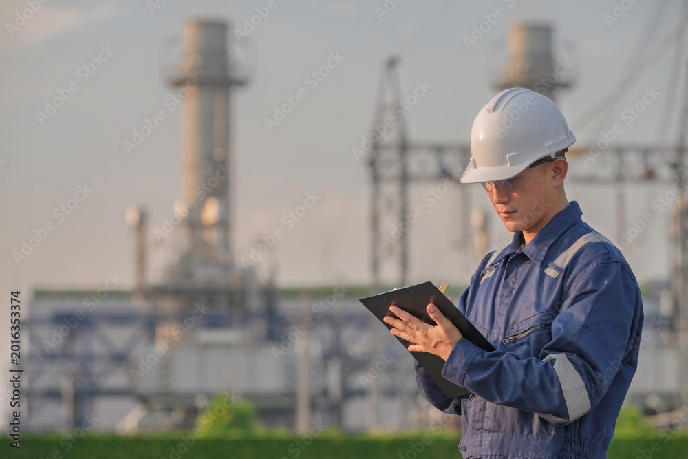 Fototapeta premium Asian male electrical engineer holding digital tablet standing at power plant station. Professional energy industry technician working on infrastructure maintenance and renewable resource project.