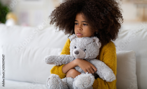 Photos Upset preteen african american girl with curly bushy hair sitting on couch alone, embracing teddy bear, looking at copy space