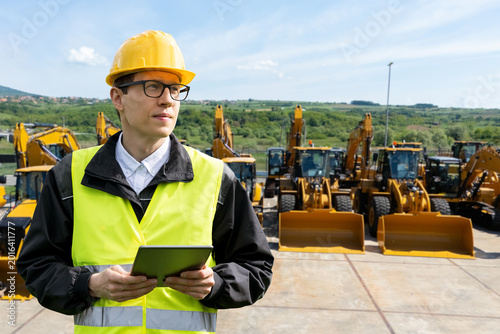 Engineer in a helmet with a digital tablet stands next to construction excavators