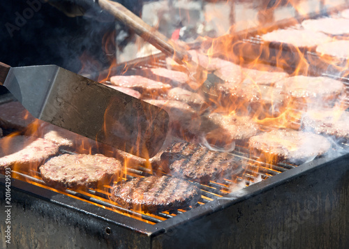 Close up on many hamburger patties, raw and cooked on an open BBQ grill outside, flames shooting up between the patties.