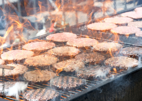 Close up on many hamburger patties, raw and cooked on an open BBQ grill outside, flames shooting up between the patties.