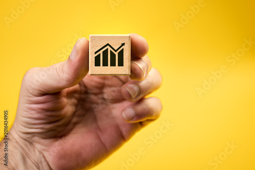 Hand holds small wooden block with simple graph symbol. Bright yellow background. Symbol suggests growth, trend, or data analysis