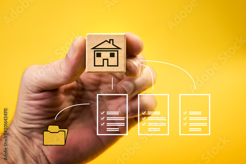 Real estate, property registration, mortgages, and data storage. Icon of a house drawn on a wooden cube held by a hand on top of documents and a folder connected by white lines.