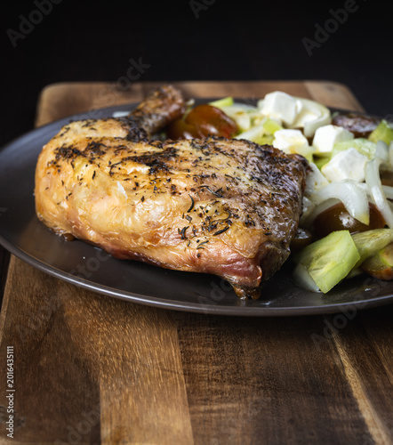 Grilled chicken thigh rests on dark plate. Golden brown skin shows crisp texture. Fresh herbs coat the meat generously. Sliced onions and cucumbers accompany it