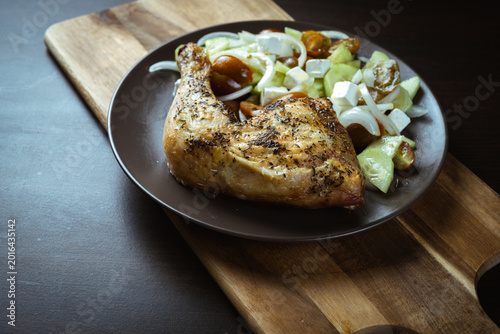 Golden chicken leg rests on dark plate. Fresh diced vegetables surround the meat. Wooden cutting board holds the dish. Soft lighting highlights texture and color