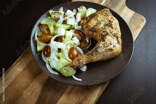 Grilled chicken leg rests beside a fresh cucumber-tomato salad. Dark plate sits on a rustic wooden cutting board. Warm tones highlight the dish's hearty appeal. Ideal for food blogs, recipe guides