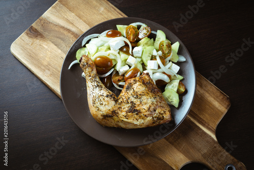Golden-brown chicken leg rests on dark plate. Crisp cucumber, diced onion, and cherry tomatoes surround it. Plated dish sits on rustic wooden cutting board