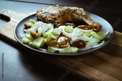 Roasted chicken leg rests beside sliced onions and cucumbers. Crispy skin gleams under warm, natural lighting. Fresh vegetables add color and crunch to the dish. Plate sits on a wooden cutting board