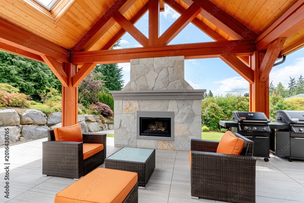 Fototapeta premium Luxurious outdoor living area featuring a stone fireplace, comfortable lounge furniture, and modern barbecue grills under a wooden gazebo