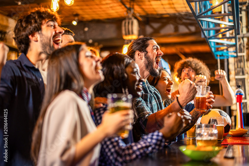 Friends cheering during soccer match in pub with beers on table.