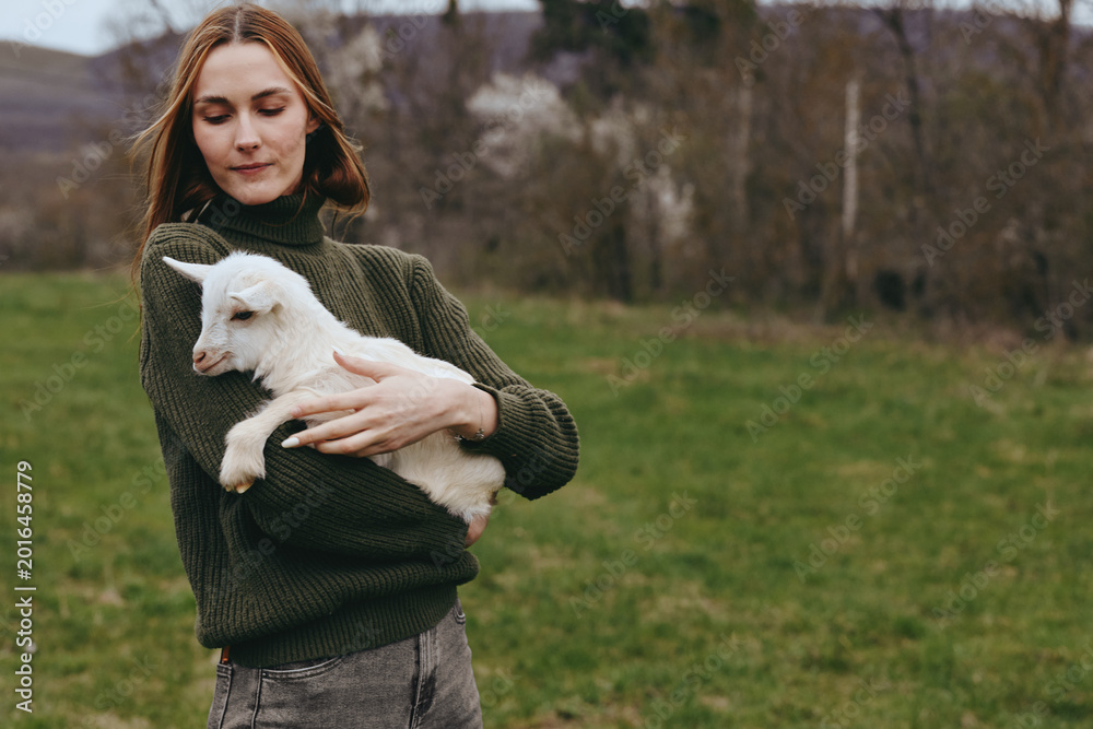 Naklejka premium Calm woman outdoors holding a goat kid close, gentle expression, olive green sweater, soft natural light, rural field backdrop, relaxed posture, warm tones, candid lifestyle moment, fresh countryside