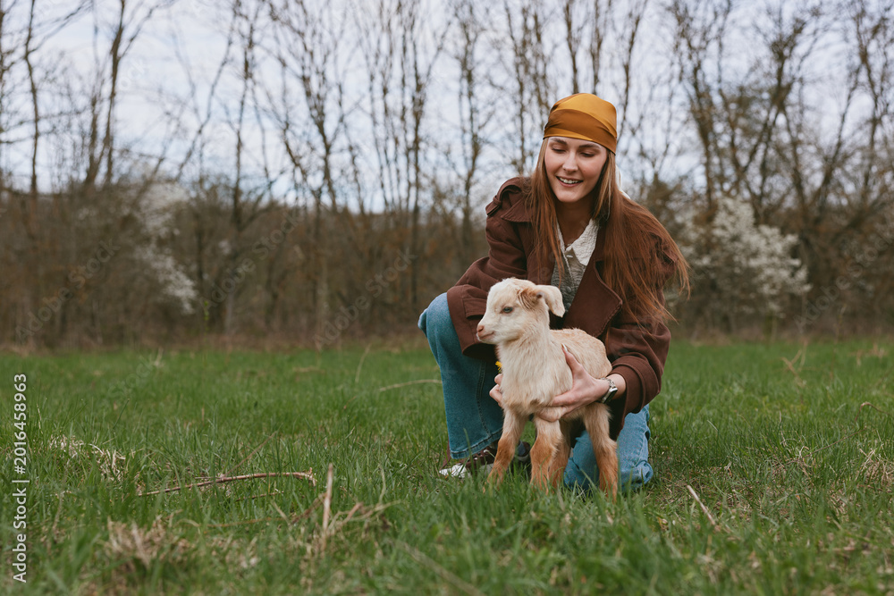 Obraz premium Cheerful woman outdoors with a goat kid, kneeling in a grassy field, warm brown jacket, light, candid lifestyle moment, gentle interaction, natural colors, tranquil countryside backdrop