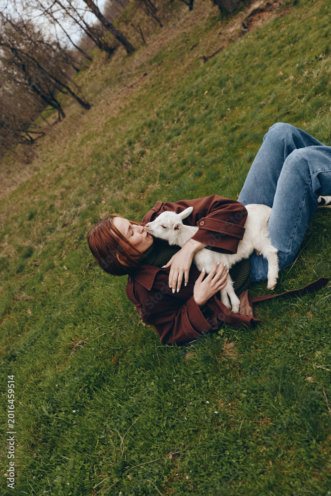 Fototapeta premium Woman lying on grass in a park, cuddling a white goat kid, enjoying a relaxed outdoor moment, soft light, natural colors, casual outfit, tranquil lifestyle scene